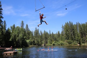 A boy in swim trunks swings on a rope over a lake at a Northern California Summer Camp, letting go mid-air. Below, children play on a floating platform, surrounded by tall pine trees and blue sky.