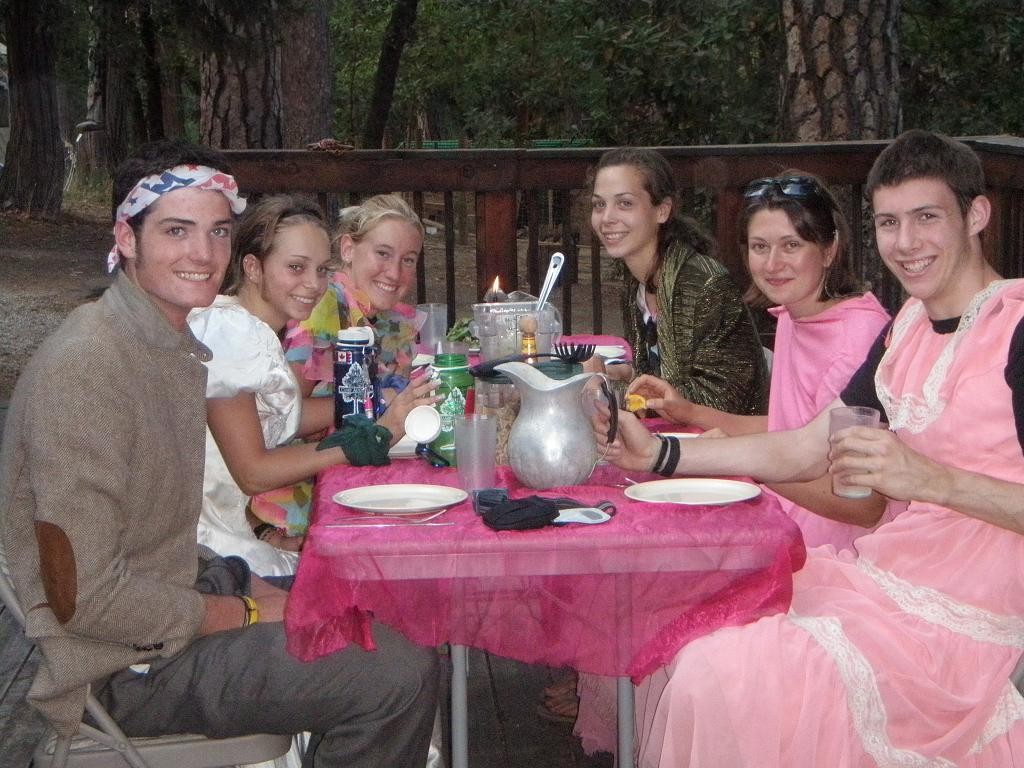 Six smiling young adults, dressed in costumes like a summer camp counselor with leis and headscarves, gather around a table outdoors. The pink cloth, surrounded by trees and a wooden fence, sets the scene for their cheerful meal.