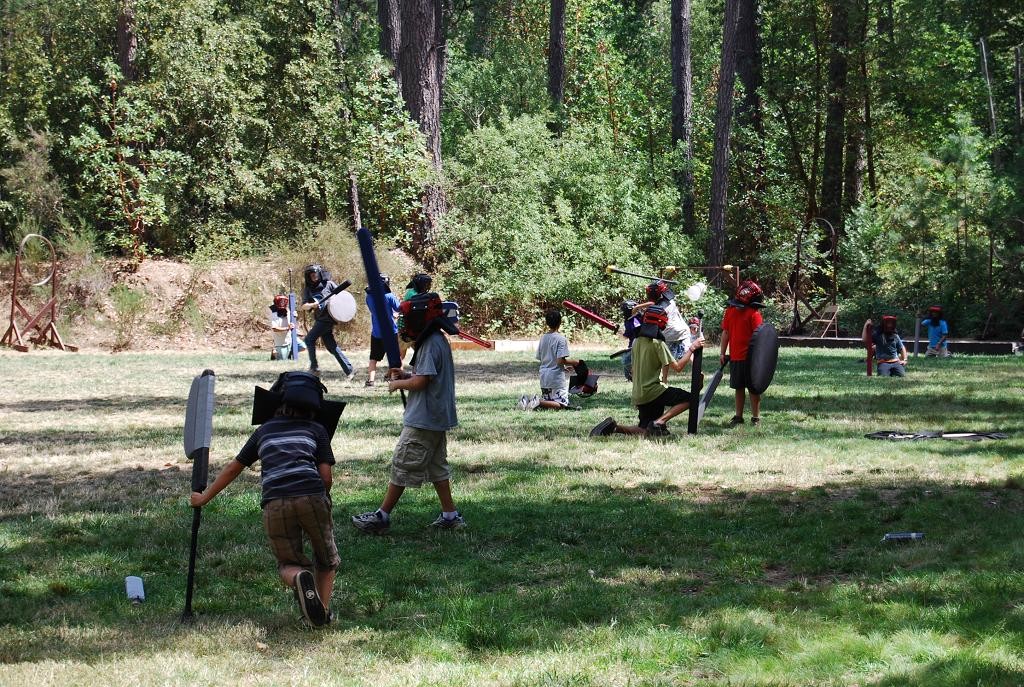 Children wearing helmets play a mock battle outdoors, using foam swords and shields on a grassy field surrounded by trees—a lively scene of summer camp activities with kids running, kneeling, and engaging in pretend combat.