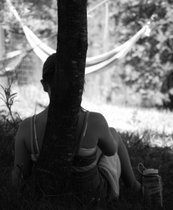 A person sits in the shade, leaning against a tree, with their back to the camera. In the background, a hammock hangs between trees in a sunlit, grassy area—capturing the peaceful side of summer camp activities. A water bottle rests nearby.