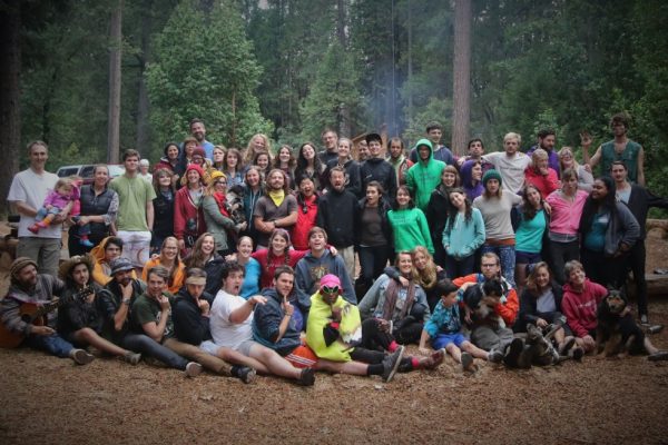 A large group of people, of various ages, pose and smile together outdoors in a forested area—some sitting, some standing—with trees and logs visible in the background, highlighting the camaraderie of a successful staff application event.