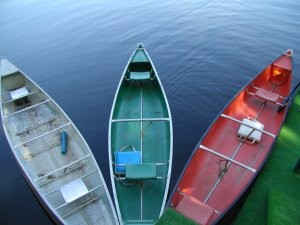 Three rowboats—one silver, one green, and one red—are docked side by side on calm, dark blue water. The scene captures the spirit of a summer camp for kids, with boat interiors and parts of the dock visible from above.