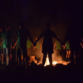 A group of people stand in a circle holding hands around a campfire at night, their faces dimly lit by the fire’s glow against the dark background.