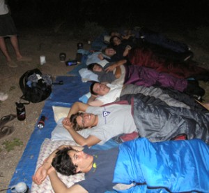 A group of people lie side by side in sleeping bags on a tarp outdoors at night, looking up and relaxing together, with backpacks and water bottles nearby—a perfect example of classic summer camp activities.
