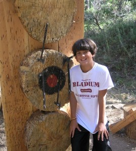A smiling boy stands next to a wooden target outdoors with three throwing knives in the bullseye, enjoying summer camp activities. He wears a white Bladium Lacrosse T-shirt and brown shorts, surrounded by trees and bushes.