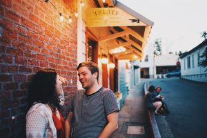 A woman and man stand laughing together outside a shop named Treats on a brick-lined street at dusk, with warm string lights overhead and people sitting further up the sidewalk.