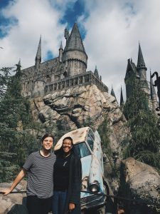 Two people stand smiling in front of a castle resembling Hogwarts, surrounded by rocky terrain and trees, with a blue, weathered car partially embedded in the rocks below the castle. The sky is partly cloudy.