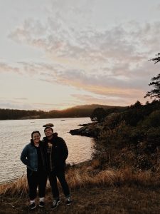 Two people stand smiling together near the edge of a calm lake at sunset, with grassy ground and bushes around them, hills and trees in the background, and a colorful sky with scattered clouds overhead.