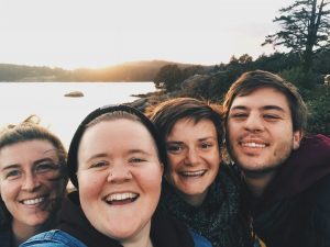 Four friends smile at the camera during sunset by a lake, with water, trees, and hills in the background. The group appears happy, enjoying time outdoors together.