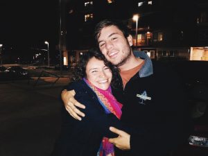 Two people are hugging and smiling warmly at the camera outside at night. The woman wears a colorful scarf and the man wears a dark jacket. Streetlights and parked cars are visible in the background.