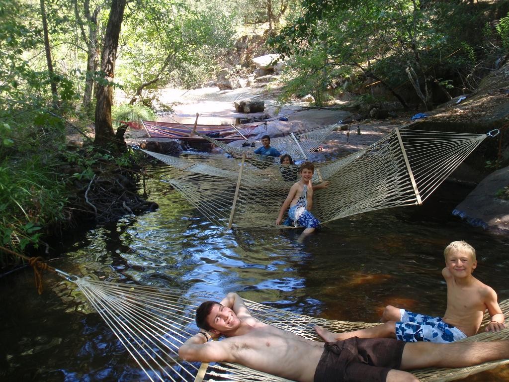 Four boys relax on hammocks suspended over a shallow creek in a wooded area, enjoying summer camp activities. Two smile at the camera while two others recline in the background, surrounded by trees and rocks.