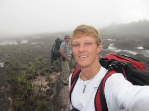 Two hikers with backpacks walk along a misty, rocky trail. The person in front, wearing a white shirt, takes a selfie while the other, in gray, stands behind surrounded by low vegetation and fog.