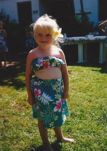 A young girl with blonde hair and a yellow flower behind her ear smiles outdoors, wearing a green floral Hawaiian dress with a cutout. She stands barefoot on the grass, with a table and other children in the background.