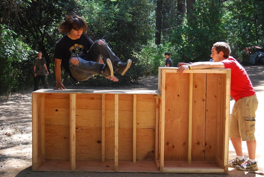 A boy leaps over a large wooden structure outdoors during family camp activities, while another boy in a red shirt holds it steady. Trees and two other people are visible in the background.
