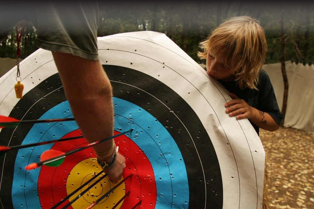 A child closely examines an archery target while an adult points to arrows clustered in the center. The setting appears to be outdoors with trees and leaves on the ground.