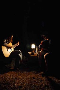 Two people play acoustic guitars in the dark, illuminated by a lantern placed on a table between them. The scene is outdoors and their faces are mostly in shadow.