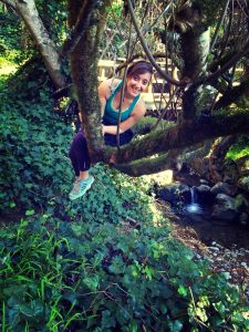 A woman in athletic wear smiles while climbing on tree branches over a small stream in a lush, green forest. Sunlight filters through the leaves, illuminating the scene.