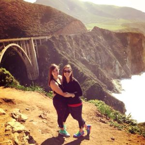 Two women smiling and hugging on a sunny cliffside trail with a large arched bridge and dramatic coastal cliffs in the background. Lush green hills and the ocean are visible under a partly cloudy sky.