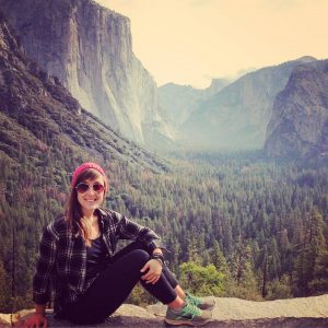 A woman in sunglasses, a red beanie, and a plaid shirt sits on a stone ledge overlooking a vast forest and dramatic granite cliffs in Yosemite National Park.