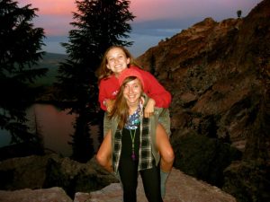 Two smiling young women outdoors; one gives the other a piggyback ride on rocky terrain at sunset, with evergreen trees, a lake, and mountains in the background.