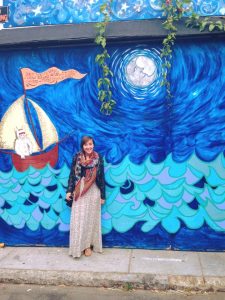 A woman in a patterned dress and colorful scarf stands smiling in front of a vibrant mural of blue ocean waves, a full moon, and a sailboat with a white creature on it. Green vines hang from above the mural.