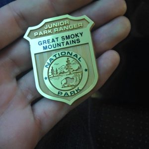 A hand holds a gold-colored badge that reads Junior Park Ranger, Great Smoky Mountains National Park with an emblem of a bear and pine trees.