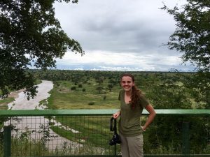 A woman stands by a green railing holding a camera, smiling with a scenic view of a river, grassy plains, and cloudy sky in the background. Trees frame the image on both sides.