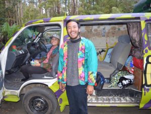 Two people smiling by a colorful, graffiti-covered van parked in a wooded area. One person sits in the driver’s seat holding a cup, while the other stands outside the van, both wearing bright jackets. Items are scattered inside the van.