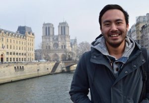 A smiling person in a gray jacket stands near a river with the Notre-Dame Cathedral and Parisian buildings in the background on a cloudy day.