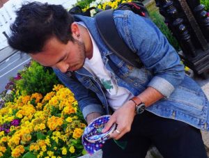 A man in a denim jacket and backpack sits by a flower bed, looking down as he holds a colorful fabric pouch in his hands. Bright yellow flowers and greenery surround him on a sunny day.