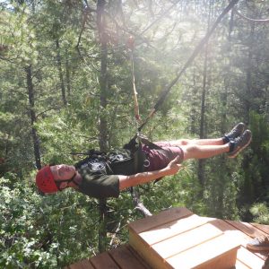 A person wearing a red helmet and harness is ziplining horizontally through a forest, above wooden platforms, with sunlight filtering through the trees in the background.