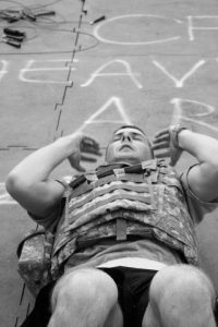 A person wearing a camouflage vest is lying on their back performing sit-ups on a gym floor with chalk writing partially visible beneath them.