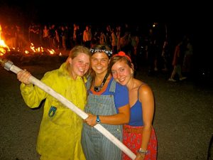 Three smiling young women pose together at night in front of a bonfire. One wears a yellow firefighter suit, another denim overalls, and the third a blue top with a red skirt. They are all holding a fire hose. A crowd is in the background.