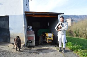 A man in a white sweater and hat stands outside a garage holding a black and white cat. A black dog walks nearby. A yellow tractor is inside the garage, with a building and hills in the background.