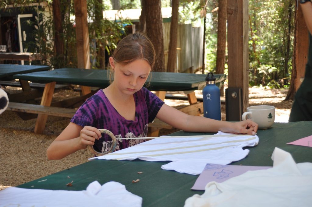 A young girl sits at an outdoor table, carefully placing masking tape on a white T-shirt for a craft project—a classic example of family camp activities. She is focused, surrounded by picnic tables, a water bottle, and trees in the background.
