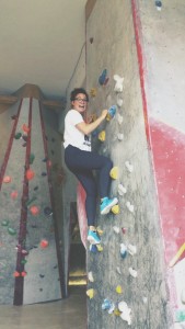 A person wearing glasses, a white t-shirt, and blue shoes is smiling while climbing an indoor rock wall with colorful holds. Another climbing wall is visible in the background.