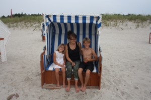A woman and two children, one girl and one boy, sit smiling together on a striped blue and white beach chair on a sandy beach, with grass and trees in the background.