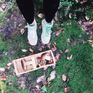 Person wearing white sneakers and black leggings standing on green mossy ground beside a wooden basket filled with freshly picked mushrooms and surrounded by fallen leaves.
