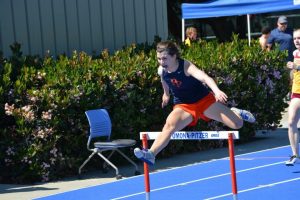 A female athlete in a navy blue and orange uniform leaps over a hurdle on a blue track during a race, with another runner approaching behind her and greenery in the background.