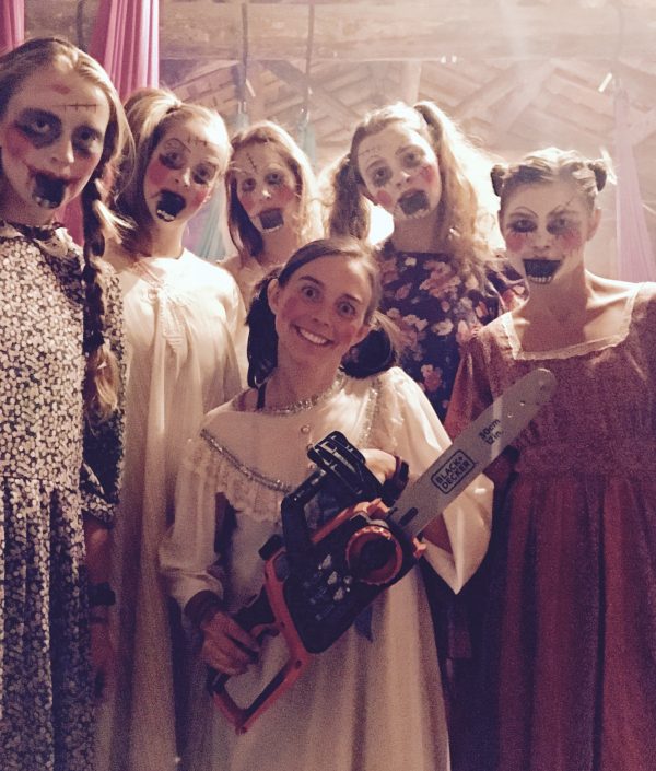 Six people in old-fashioned dresses with creepy clown-like makeup and black-painted mouths stand together indoors. One person in the front smiles while holding a toy chainsaw, while the rest stare with eerie expressions.