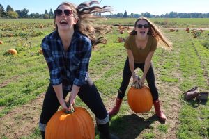 Two young women outdoors in a pumpkin patch, each lifting a large pumpkin while smiling and laughing. One wears a plaid shirt and sunglasses, the other a green shirt and red boots. The field is sunny with scattered pumpkins.