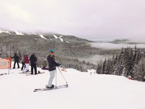A skier stands smiling on a snowy slope with ski poles, surrounded by other skiers. Snow-covered trees and distant mountains are visible under a cloudy sky.