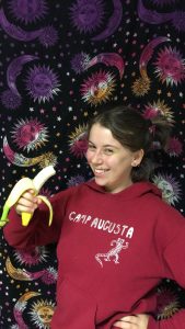 A smiling young woman in a red Camp Augusta hoodie holds a peeled banana in front of a colorful tapestry decorated with suns, moons, and stars.