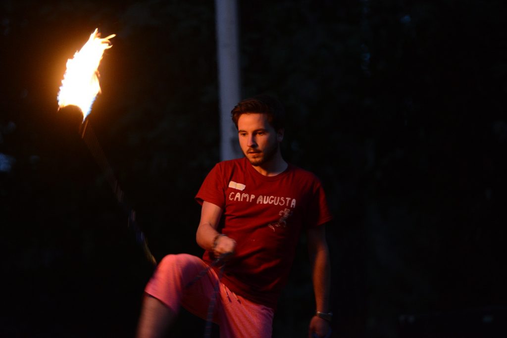 A person wearing a red Camp Augusta shirt and pink shorts is spinning a flaming torch outdoors at night, showcasing one of the exciting family camp activities, with a focused expression on their face.