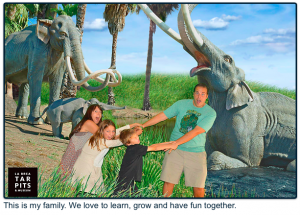 A family of four pretends to be pulled away by a woolly mammoth statue at the La Brea Tar Pits Museum, with playful expressions and green grass in the background.