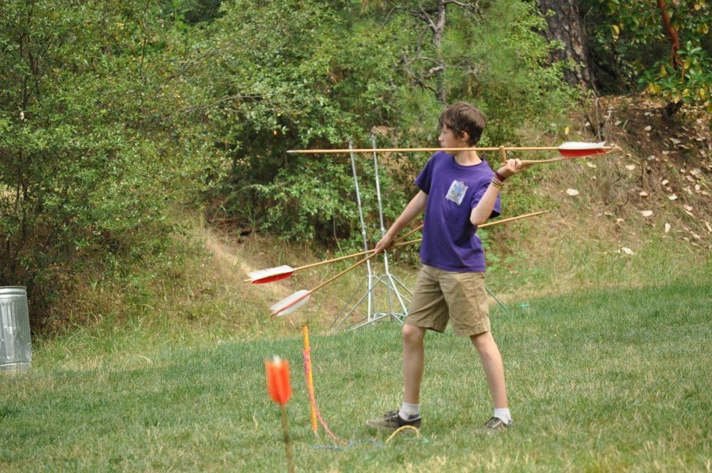 A boy in a purple shirt and khaki shorts stands on grass, holding three large foam javelins—a perfect example of family camp activities. He is about to throw one, with trees and a metal target stand in the background.