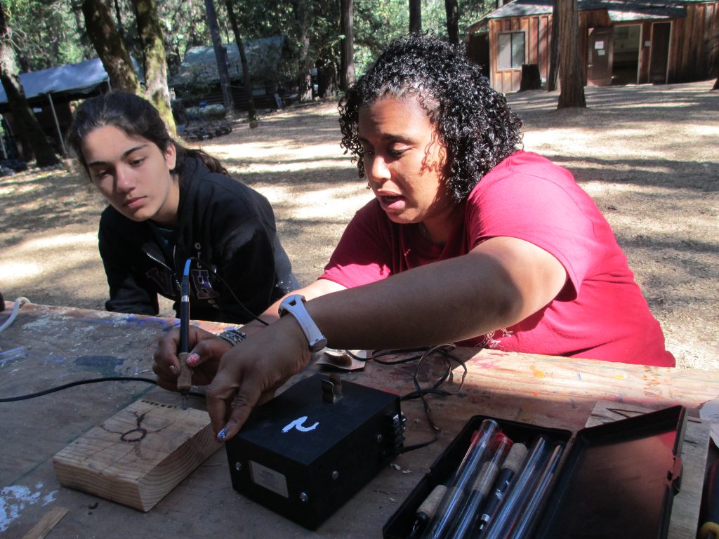 Two women sit at an outdoor table surrounded by trees and cabins, engaging in family camp activities. One woman, in a red shirt, uses a woodburning tool on wood as the other observes closely. Art supplies are scattered across the table.