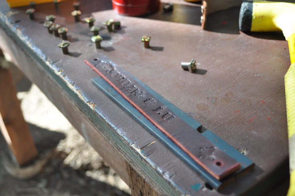 A leather strap embossed with the text JORDAN LU lies on a wooden workbench surrounded by metal rivets and a yellow tool—capturing the spirit of hands-on family camp activities as sunlight casts shadows across the surface.