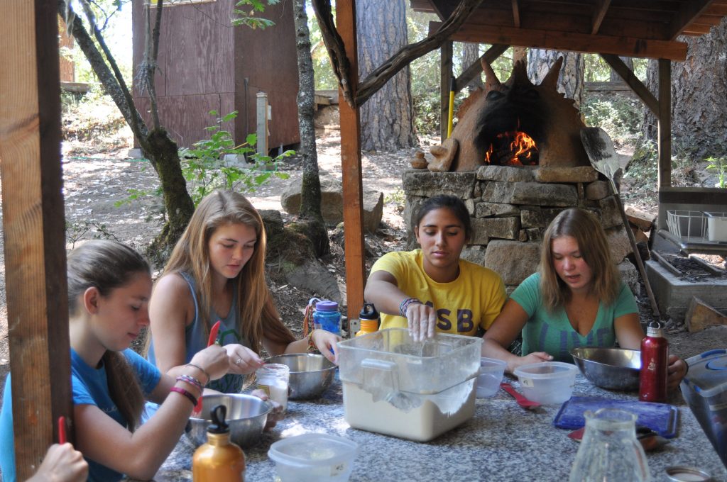 Four teenage girls sit around an outdoor table preparing food, mixing ingredients in bowls. A wood-fired oven is in the background, surrounded by trees and rustic wooden structures.