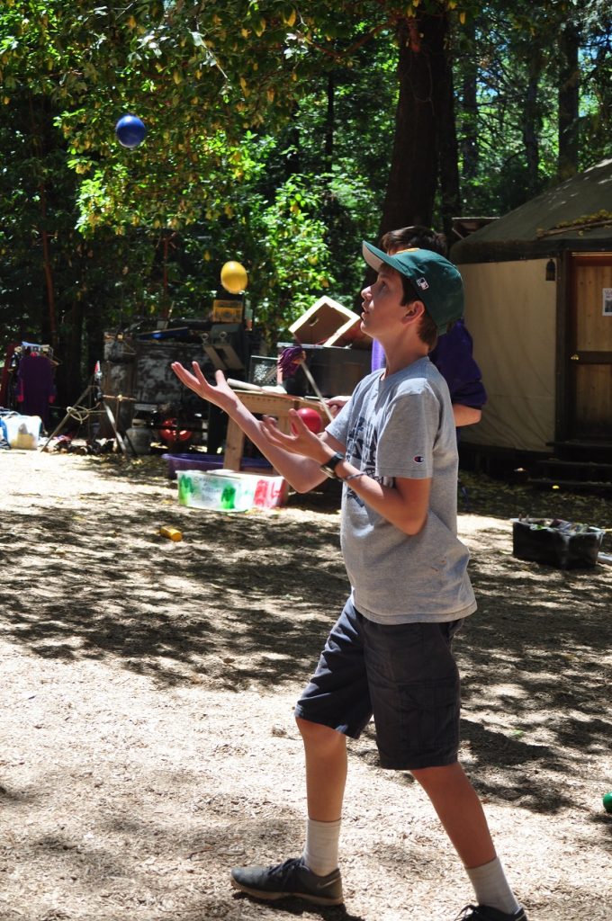 A boy in a gray shirt and shorts is juggling three colorful balls outdoors near a tent, enjoying family camp activities amid trees, scattered objects, and sunlight filtering through the leaves.